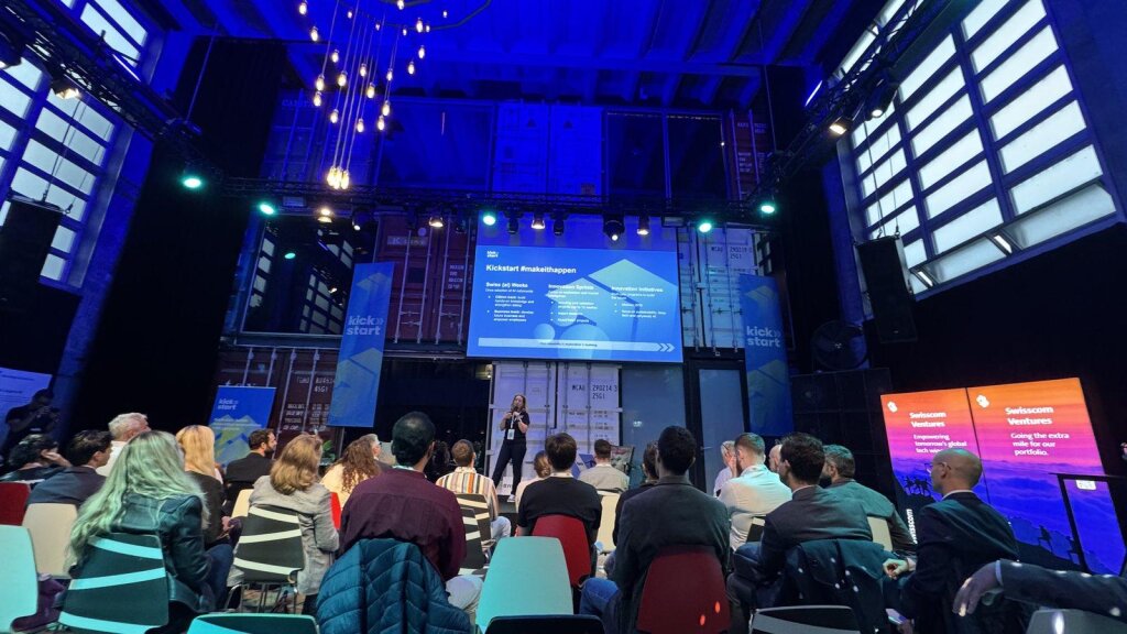 A packed room at Kraftwerk Zürich watches a Kickstart speaker present Swiss {ai} Weeks and the Innovation Sprints during Forward Days, with Swisscom Ventures banners in the foreground.